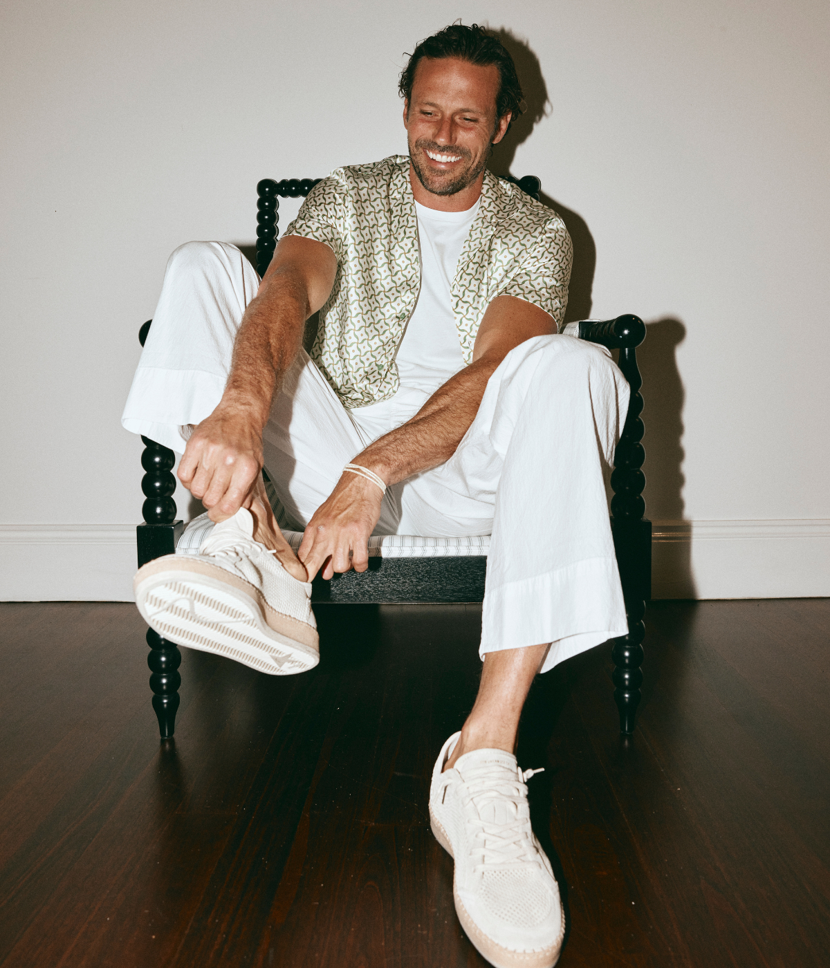 Man sitting on a chair wearing a patterned shirt and white pants, tying his white sneakers.