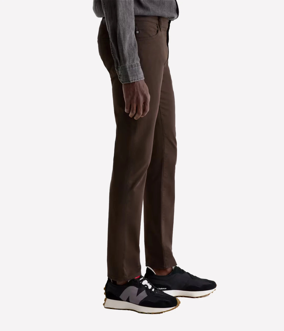 A side profile view of a person wearing slim-fit brown trousers, a gray long-sleeve shirt, and black New Balance sneakers against a plain light gray background.