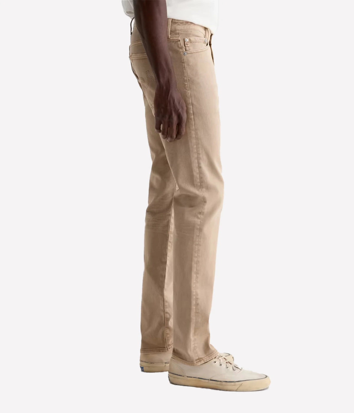 A side profile view of a person wearing beige slim-fit jeans and off-white canvas sneakers against a plain light gray background.