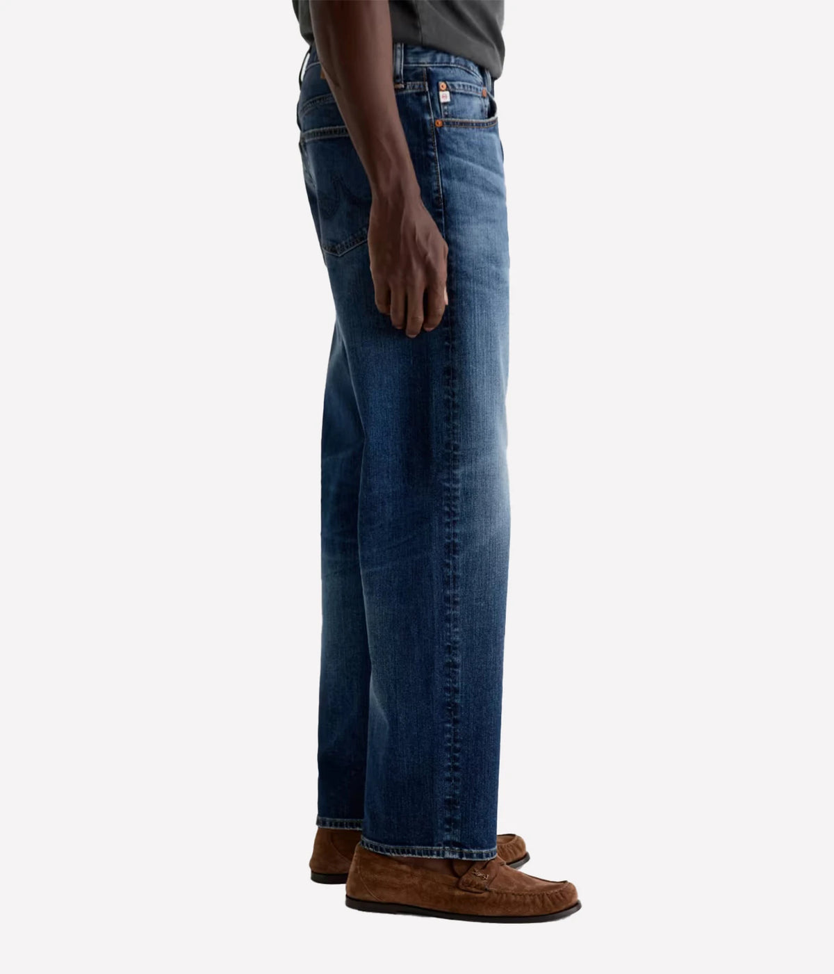 A side profile view of a person wearing medium wash blue jeans and brown suede loafers, shown from the waist down against a light gray background.