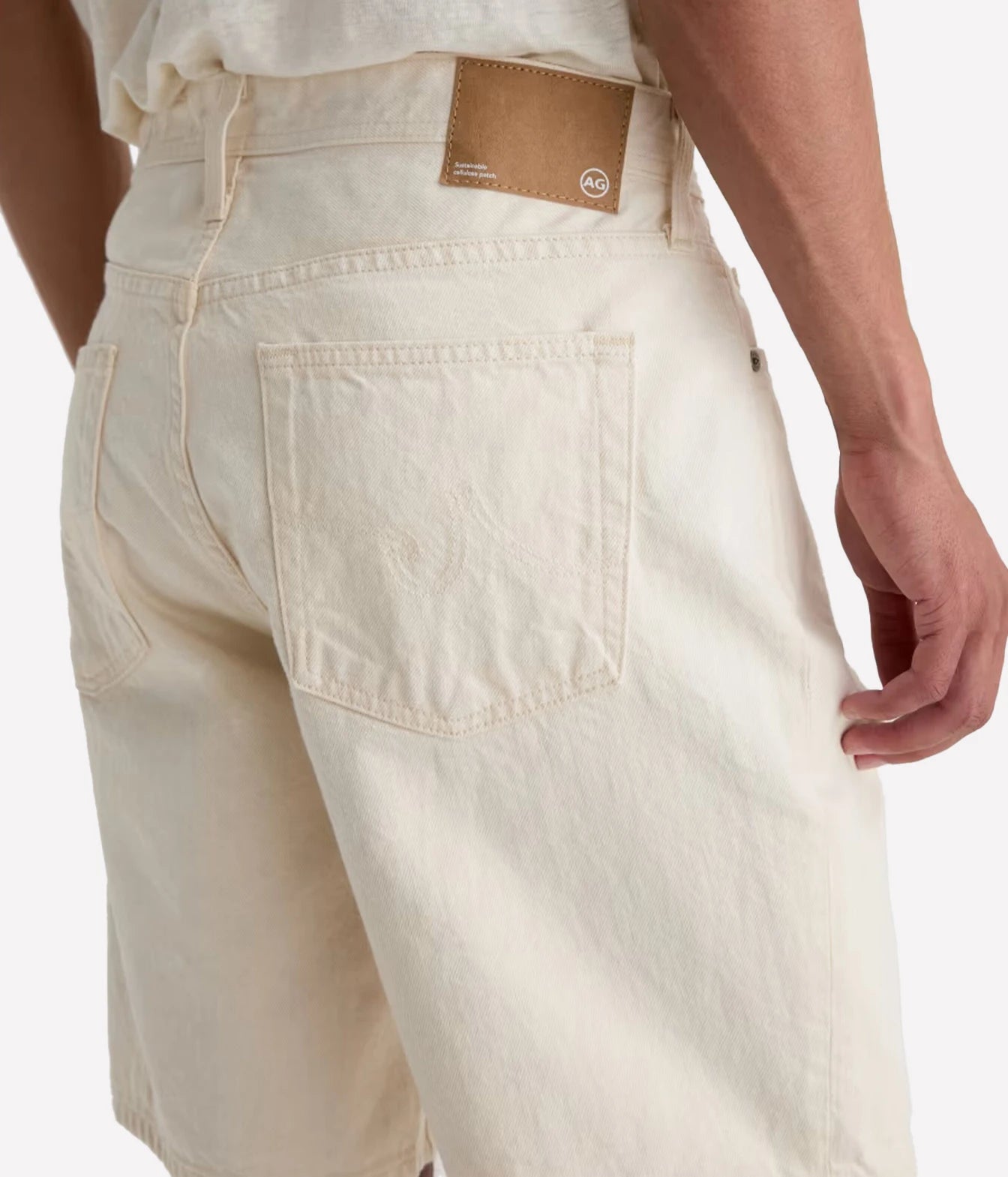 Close-up back view of a person wearing off-white denim shorts with a tan leather brand patch at the waistband, shown against a light gray studio background.