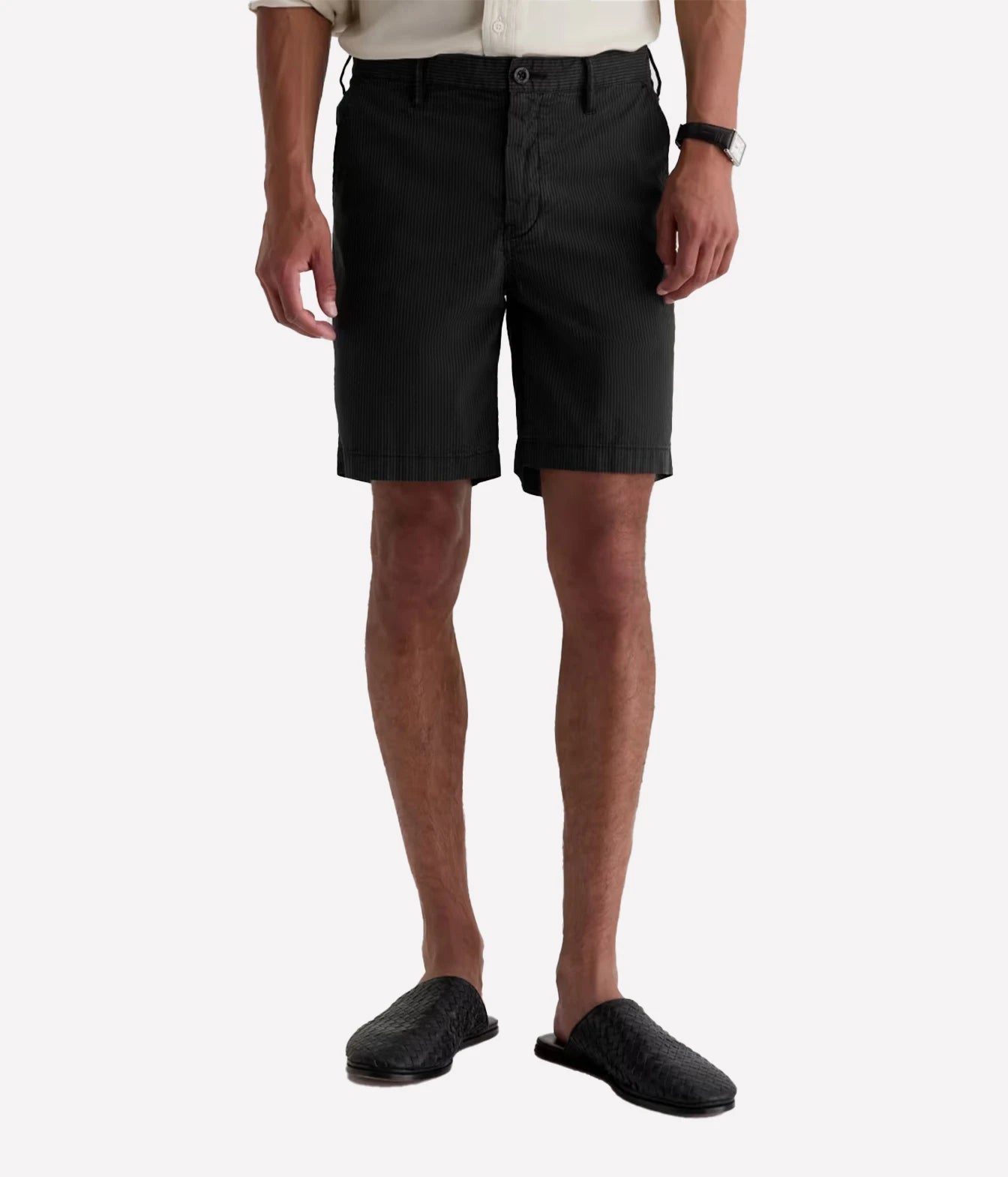A model from the waist down wearing black chino shorts, a light beige shirt, and black slip-on shoes against a light gray background.