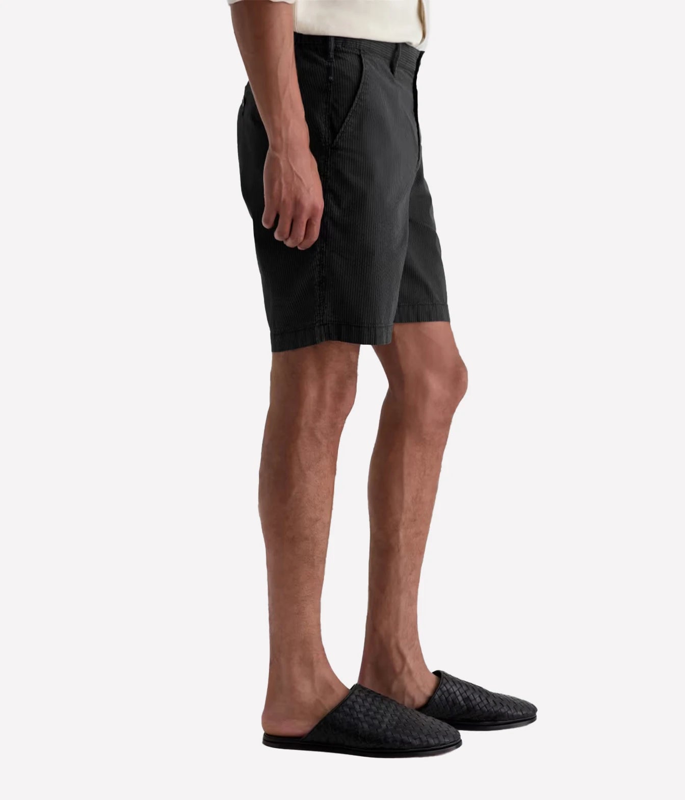 A side-profile view of a person wearing dark charcoal gray shorts, a cream shirt, and black woven slip-on shoes against a light gray background.