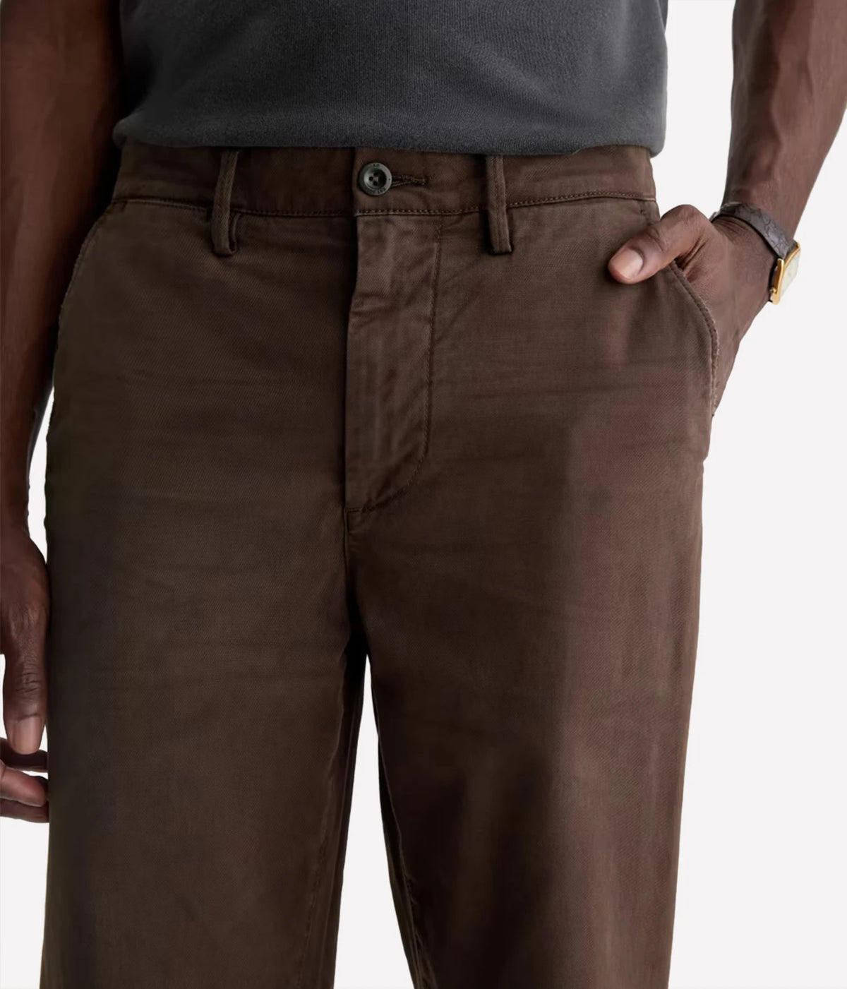 A close-up of a person wearing brown trousers, with the hands and torso visible against a plain light background.