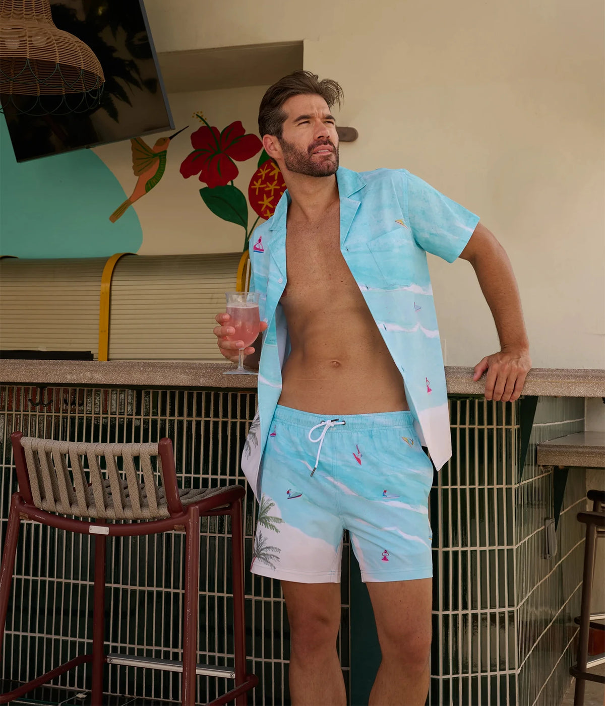 A man wearing light blue tropical print swim trunks and a matching open short-sleeve shirt stands at a bar counter holding a pink drink.