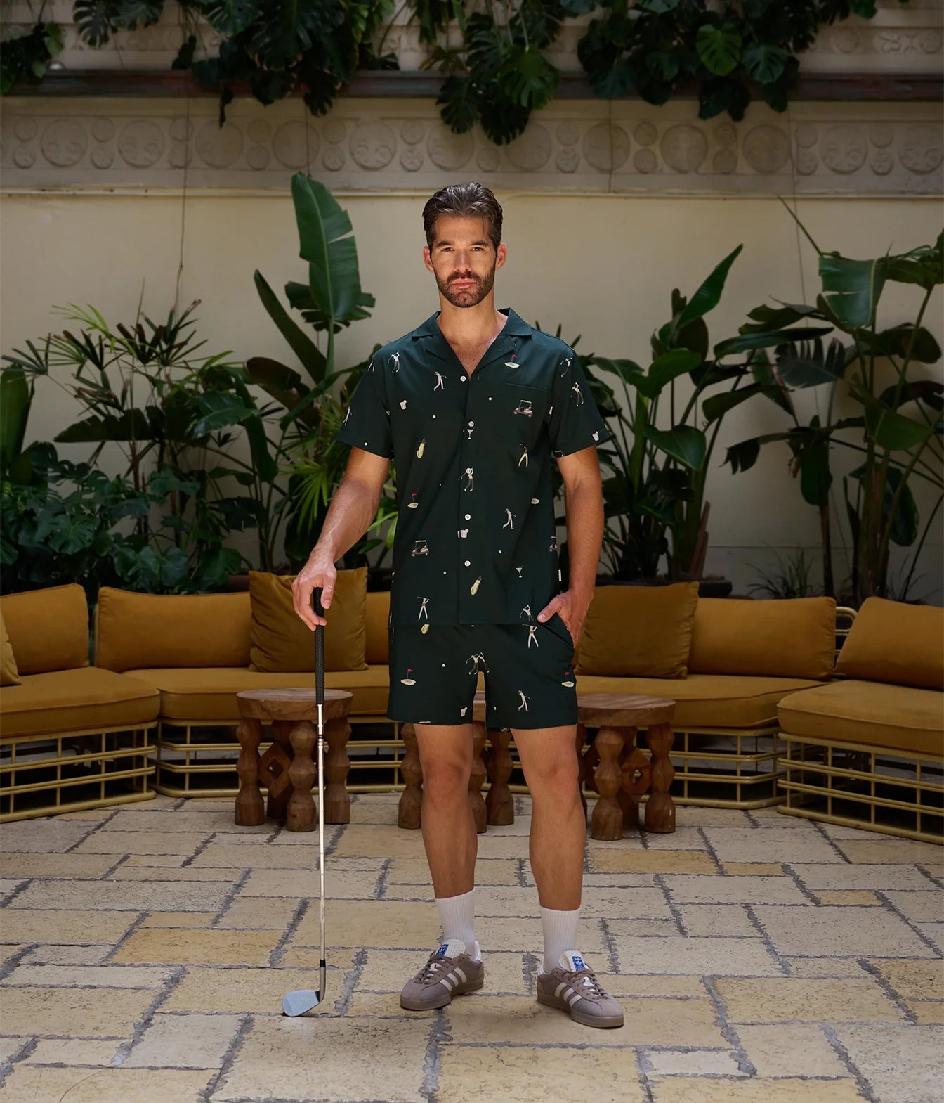 A man stands in a tropical patio lounge setting wearing a dark green short-sleeve button-up shirt and matching shorts with a white tropical print. He is holding a golf club and wearing white socks and brown shoes.