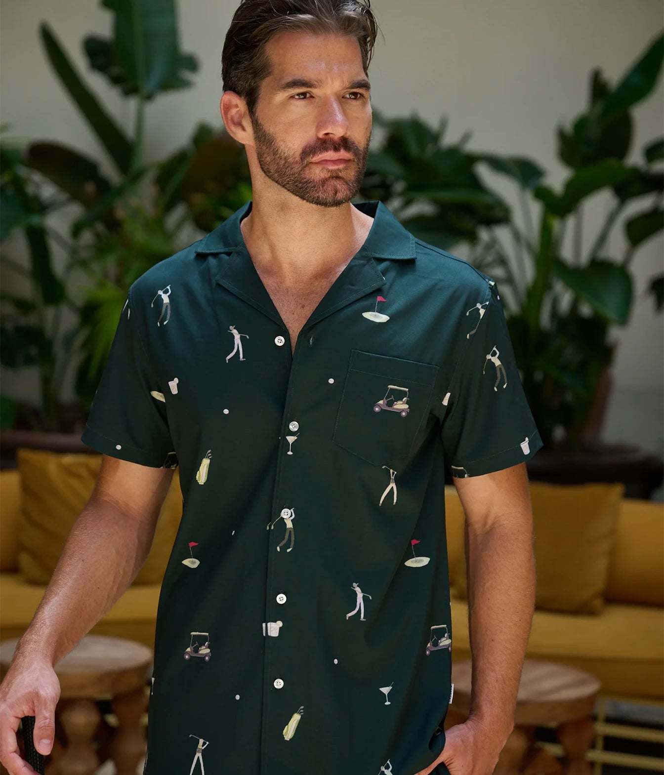 A man wearing a dark green short-sleeve button-up shirt with a golf-themed print, standing in a lifestyle setting with plants and a yellow sofa in the background.