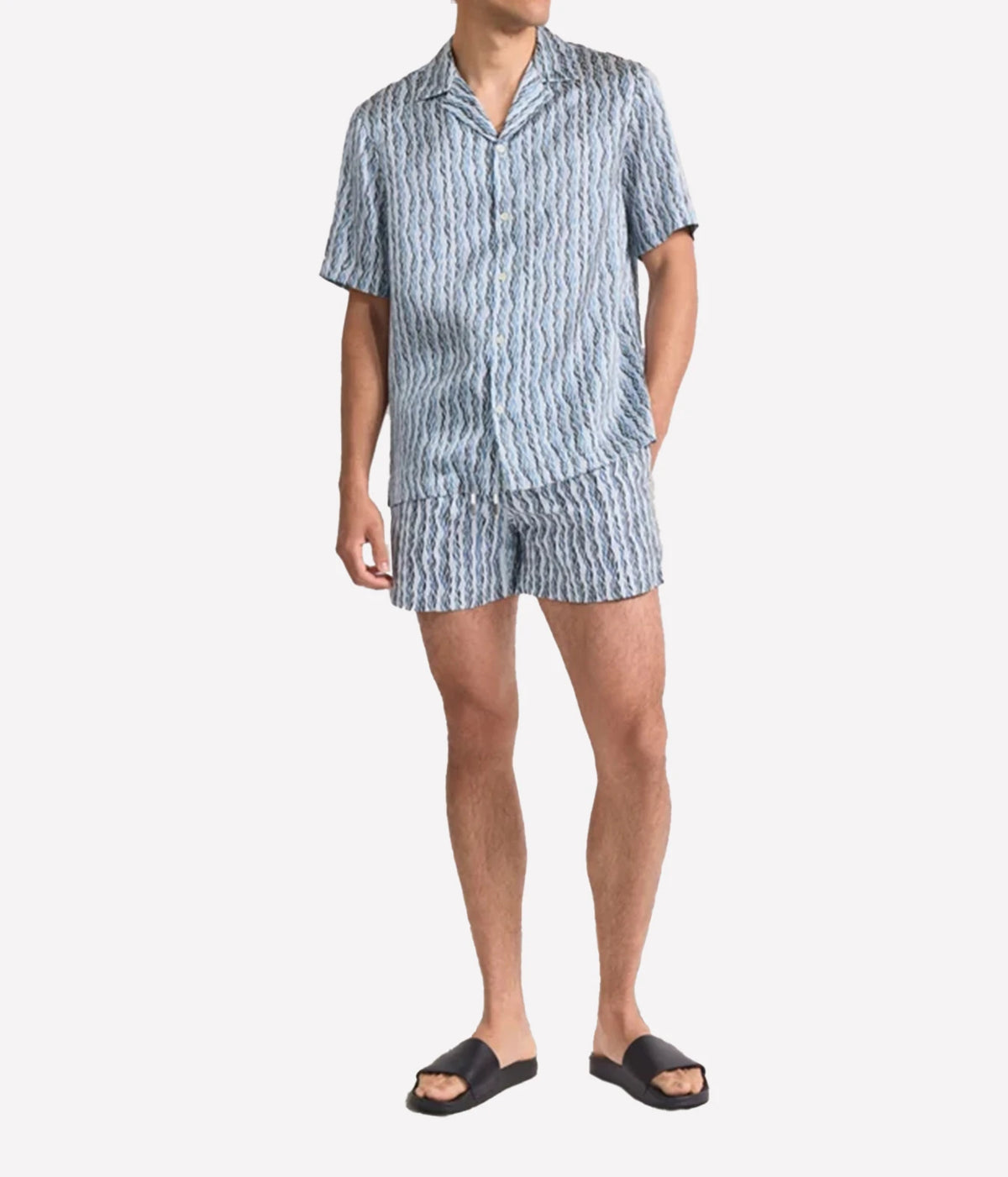 A man modeling a matching Frescobol Carioca Roberto Short Sleeve Silk Shirt in Denim Blue and shorts set with a vertical wavy stripe pattern in blue and white, paired with black slide sandals, against a light grey background.