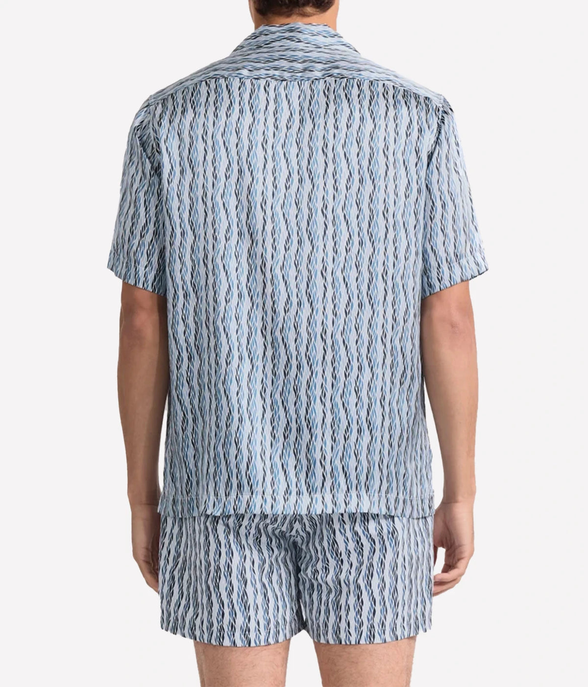 A man viewed from the back wearing a matching Frescobol Carioca Roberto Short Sleeve Silk Shirt in Denim Blue and shorts set in a light blue and white wavy striped pattern, against a plain light grey background.