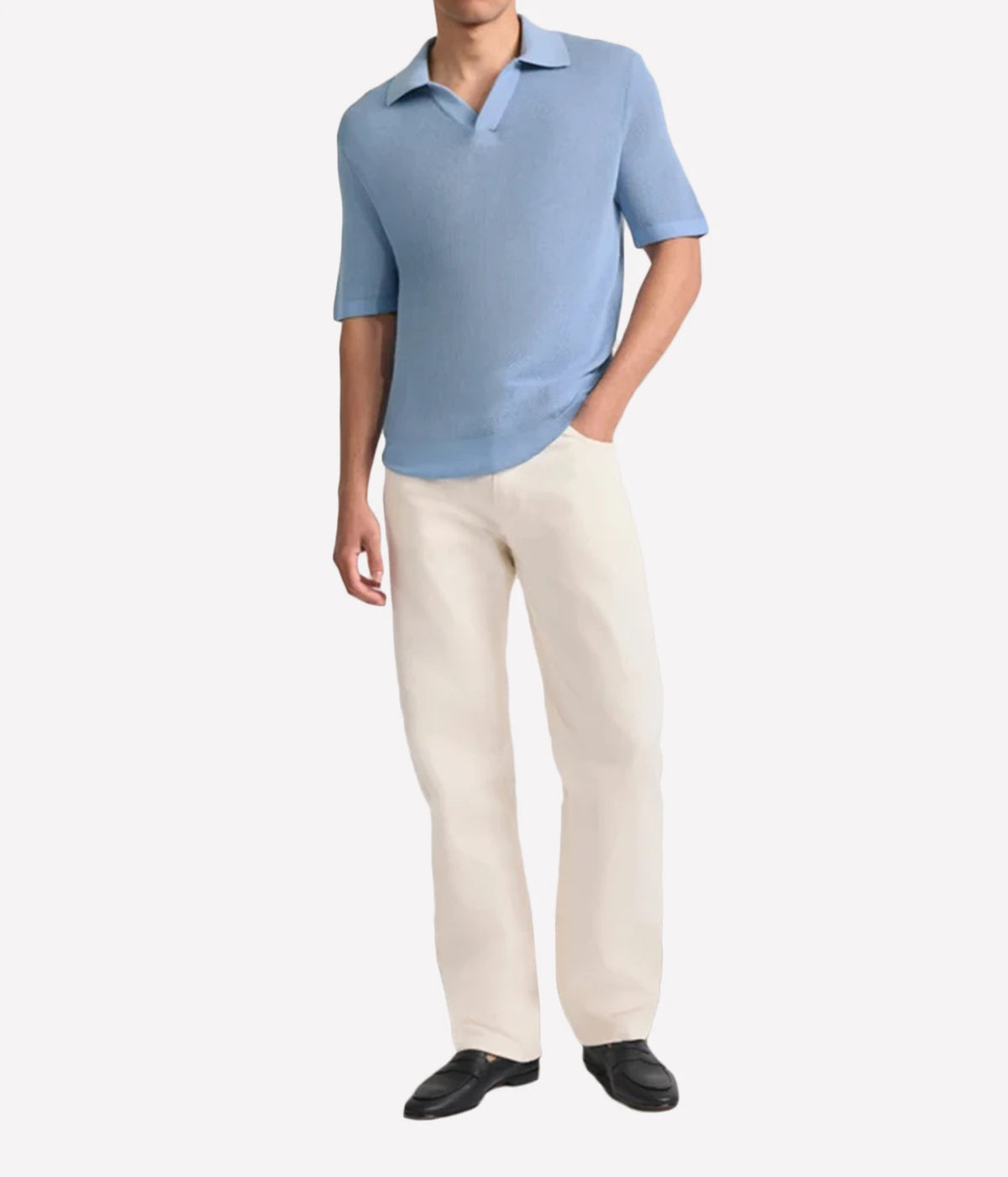 A man wearing a Frescobol Carioca Rino Short Sleeve Cotton Polo in Light Denim Blue and off-white trousers, standing against a plain light grey background.