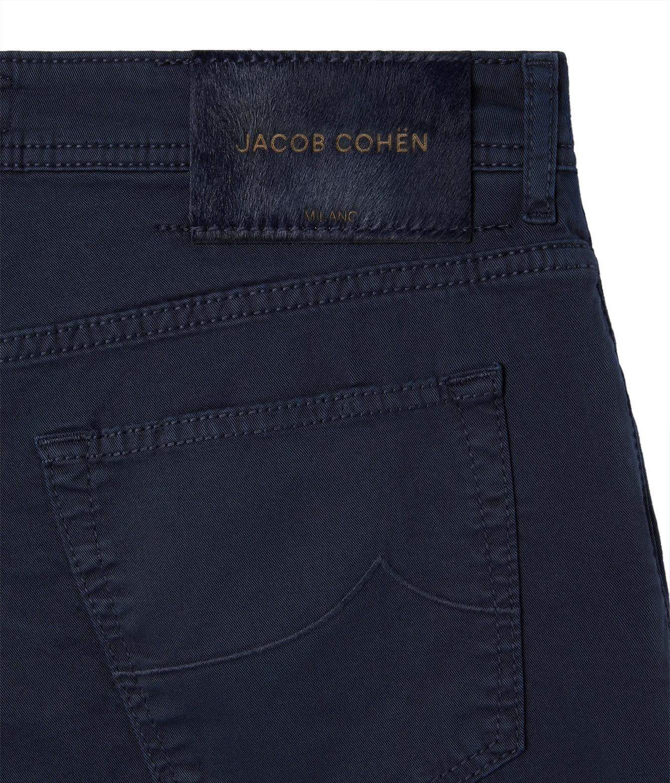 Close-up of the back waistband and pocket of dark navy blue jeans by Jacob Cohen, showing a branded leather patch and gold-tone embroidery on a white background.