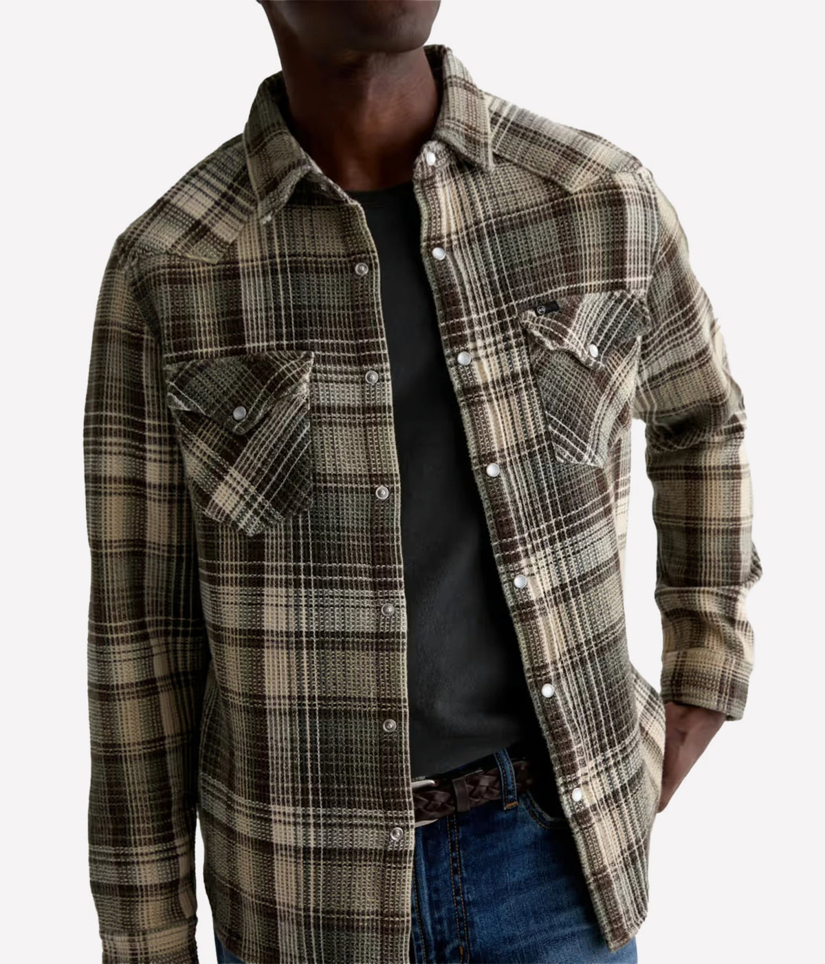 A man wearing a brown and beige plaid western-style flannel shirt over a black t-shirt and blue jeans, shown from the neck down against a light gray background.