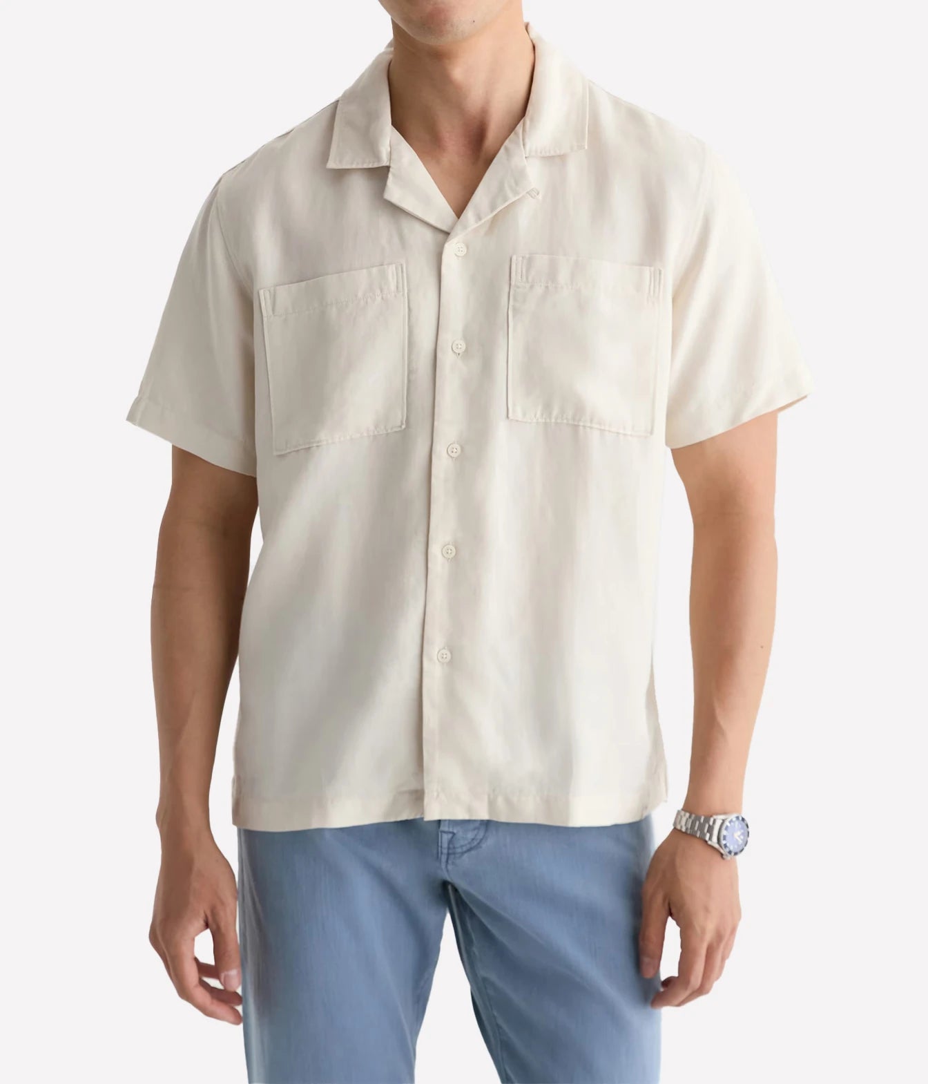 A man wearing a short-sleeve button-up shirt in a light cream or off-white color, paired with light blue pants, against a plain light gray background.
