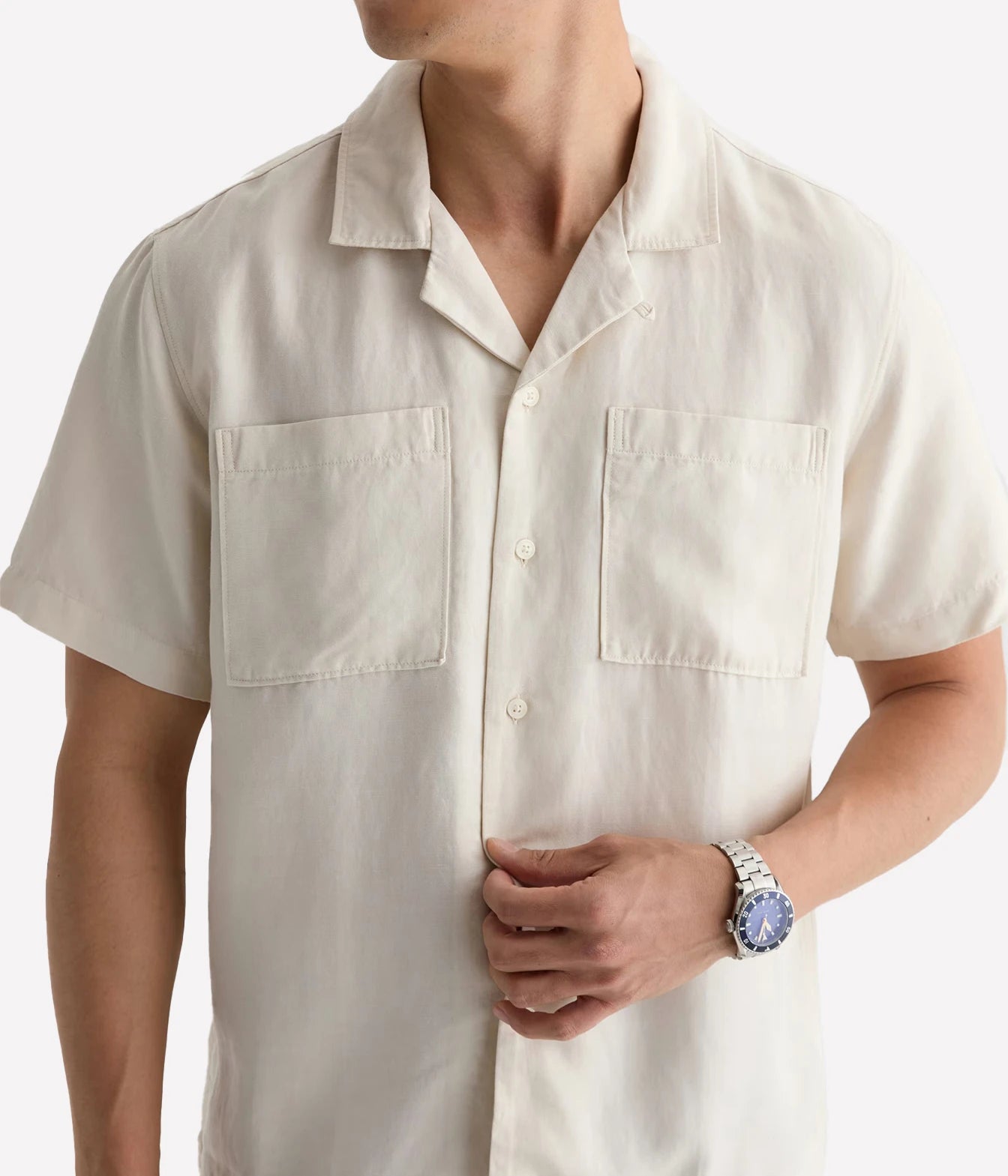 A man wearing a short-sleeve cream-colored camp collar button-up shirt with two chest pockets, shown from the neck down against a light gray background.
