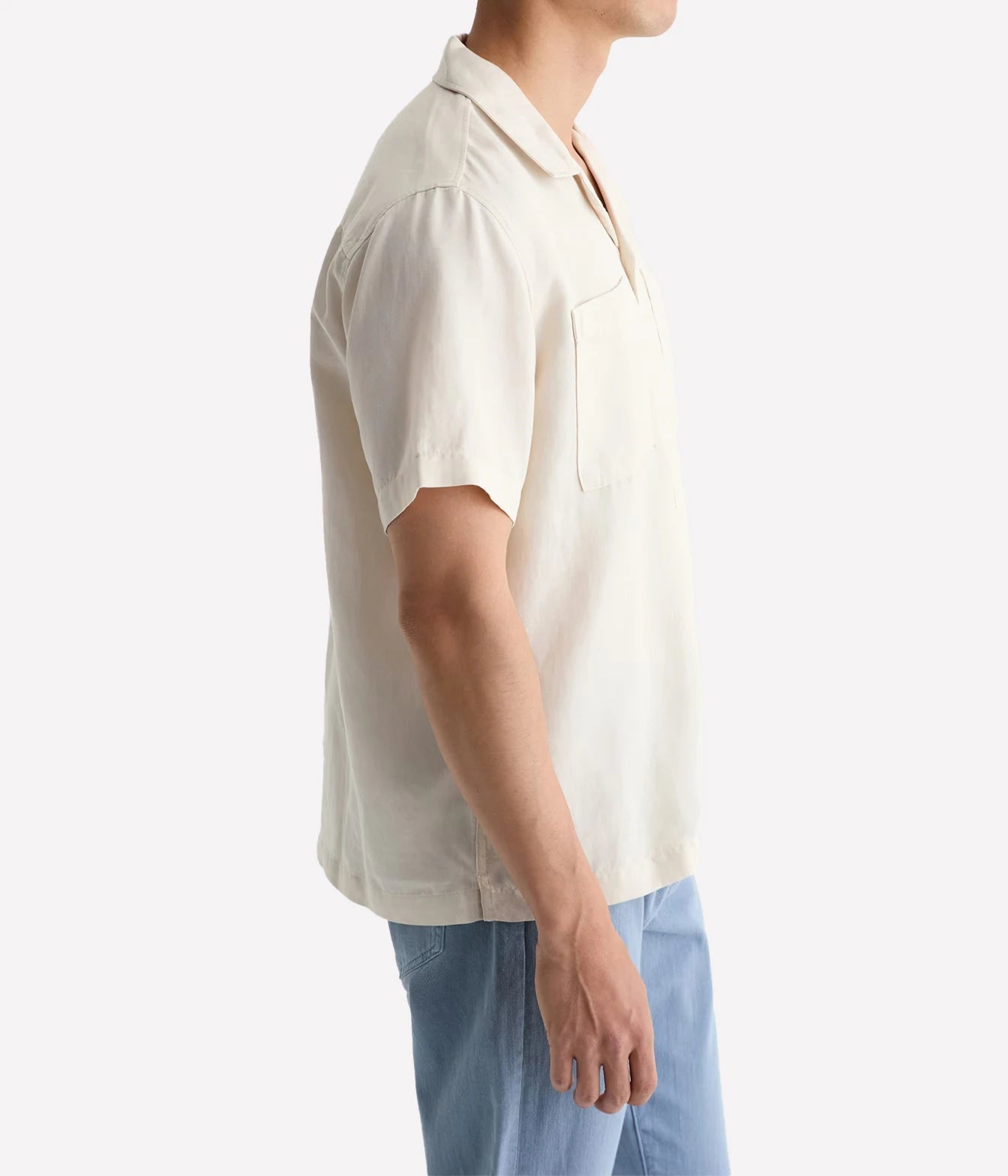 A man shown in side profile wearing a short-sleeve cream-colored linen shirt and light blue jeans against a plain white background.