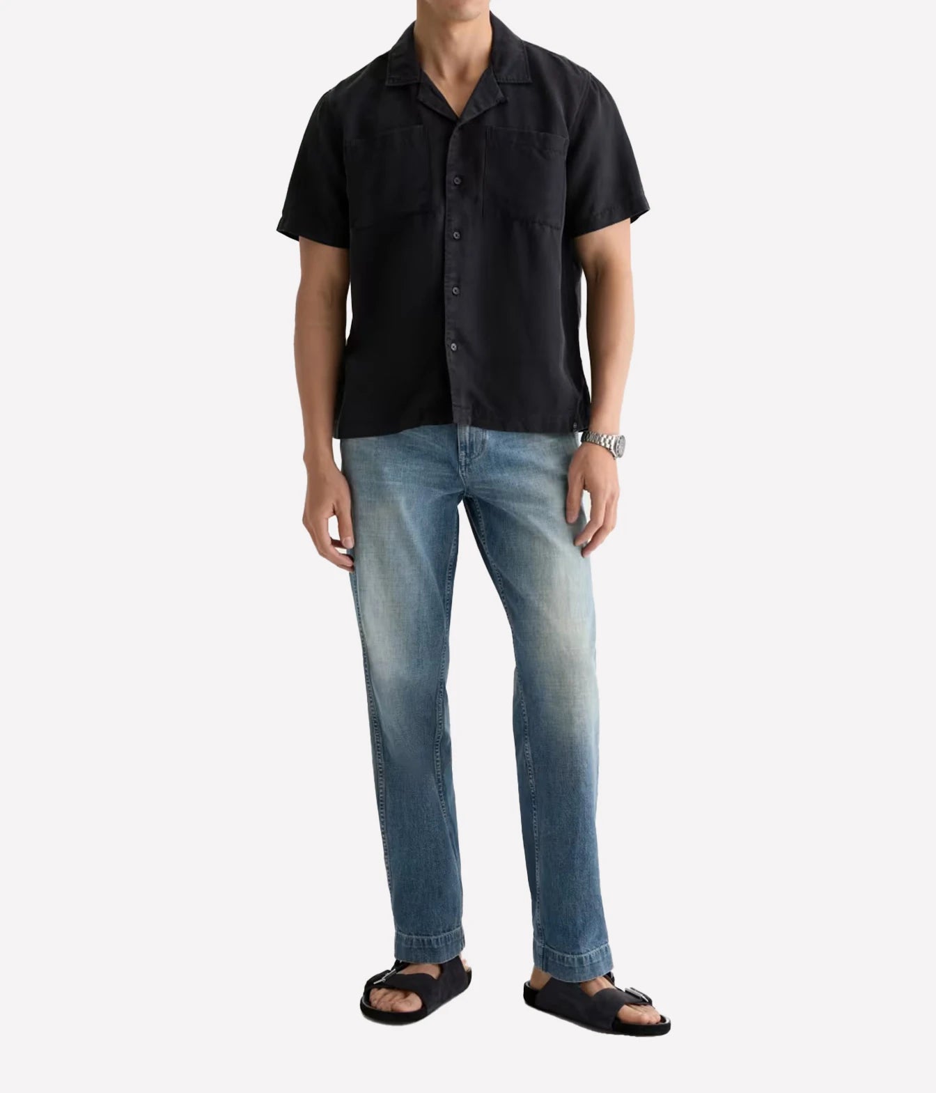 A man wearing a black short-sleeve button-up shirt, blue jeans, and black slide sandals, standing against a light gray background.
