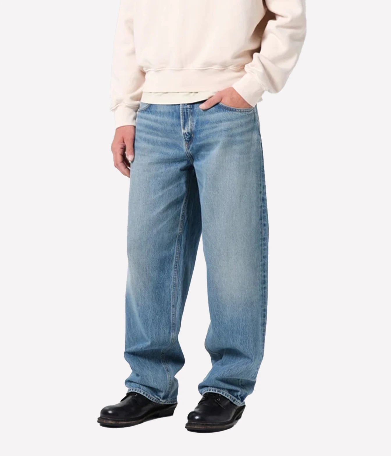 A person wearing light blue relaxed-fit jeans, a cream sweatshirt, and black shoes, shown from the chest down against a plain light gray studio background.