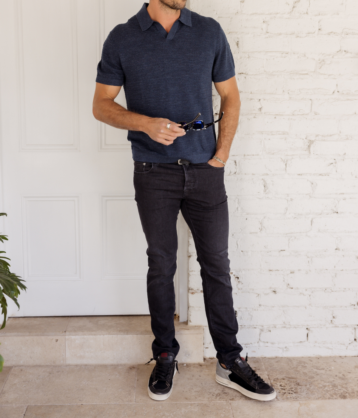 Man wearing a dark blue polo shirt and black jeans standing against a white brick wall.
