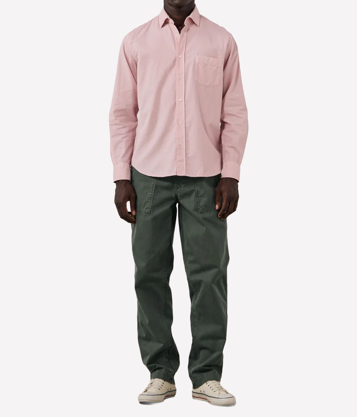 A model wearing a light pink long-sleeve button-down shirt, dark olive green pants, and beige sneakers, shown from the neck down against a plain white background.