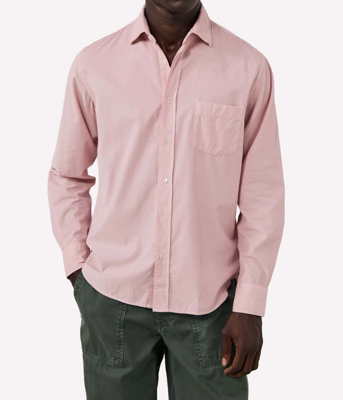 A man wearing a light pink long-sleeve button-down shirt with a chest pocket, paired with dark green pants, against a plain light grey background.