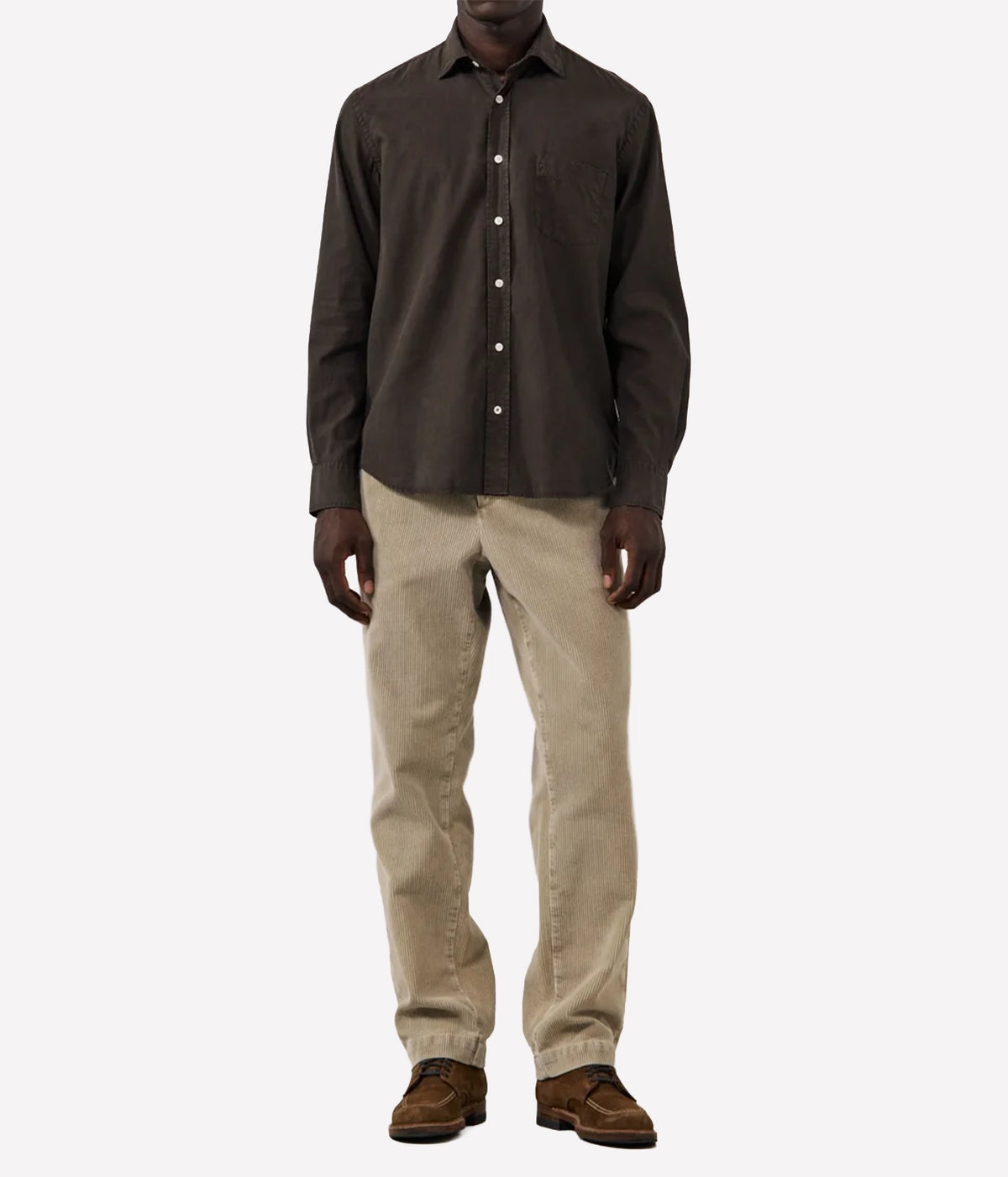 A man wearing a dark brown long-sleeve button-down shirt, beige trousers, and brown suede shoes, standing against a light grey background.
