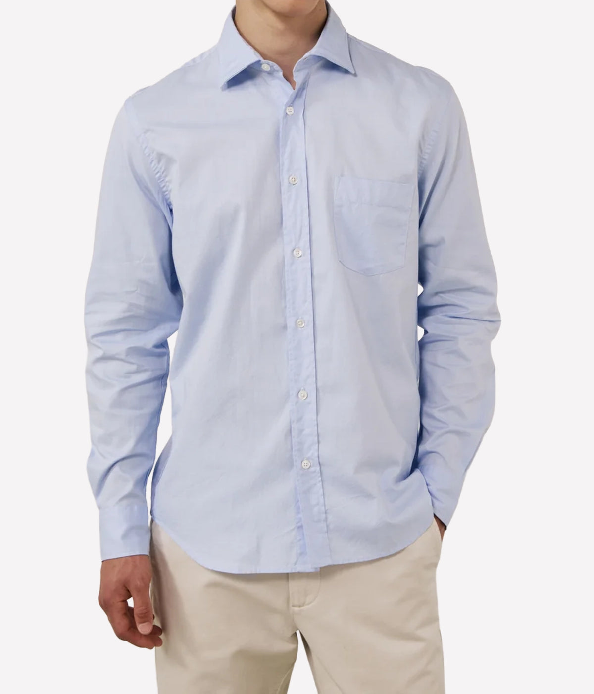 A man wearing a light blue long-sleeve button-down shirt with a chest pocket, paired with beige trousers, shown against a plain light grey background.