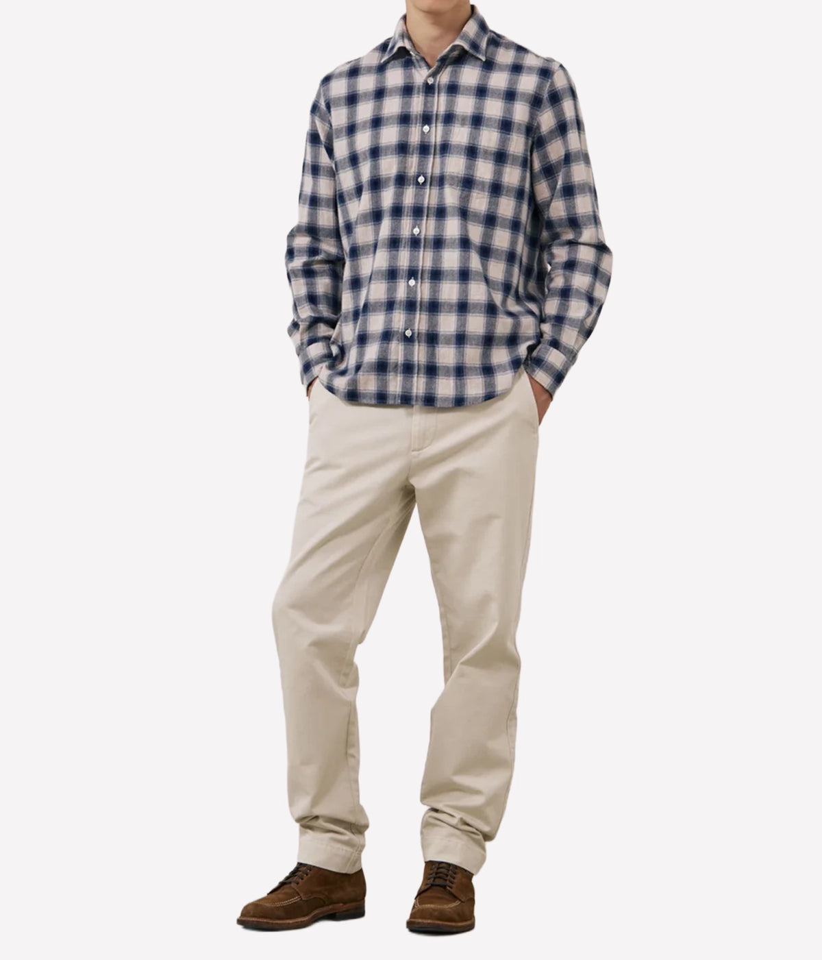 A man wearing a blue and white checkered long-sleeve button-down shirt, beige trousers, and brown shoes, standing against a light grey background.