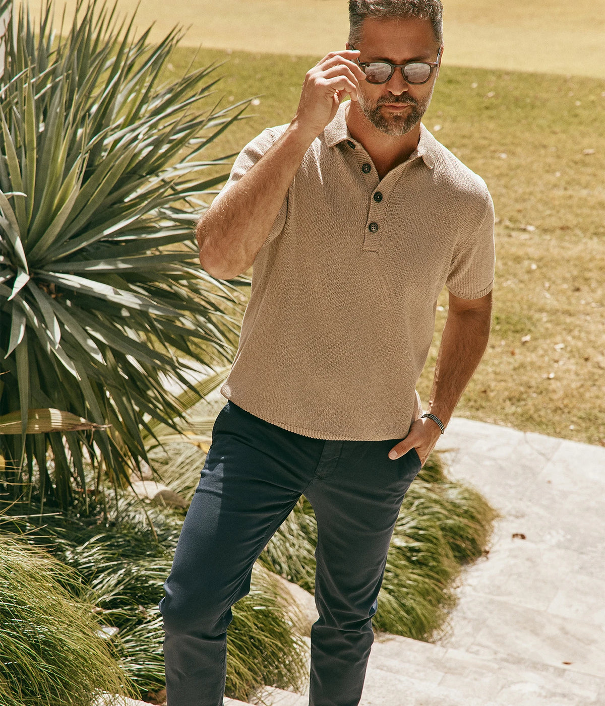 Model wearing the Eleventy Polo Sweater in Taupe with dark-wash jeans, adjusting his sunglasses with one hand, front view