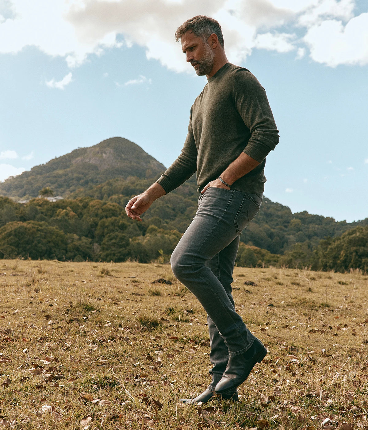Man wearing Avant Toi Hand Painted Light Cashmere Round Neck Pullover in Moss with jeans, walking through a field