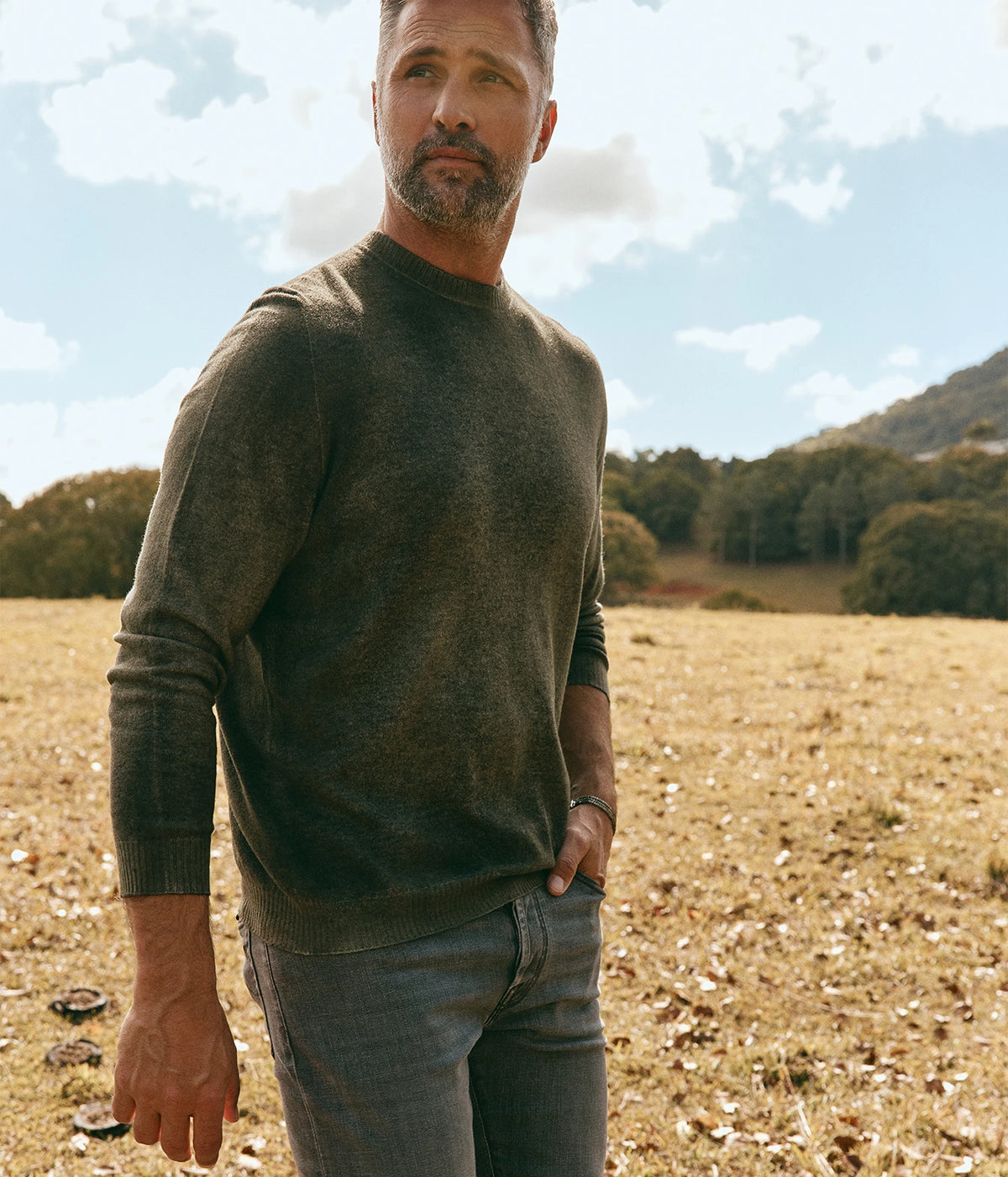 Man wearing Avant Toi Hand Painted Light Cashmere Round Neck Pullover in Moss outdoors in a field, three-quarter view