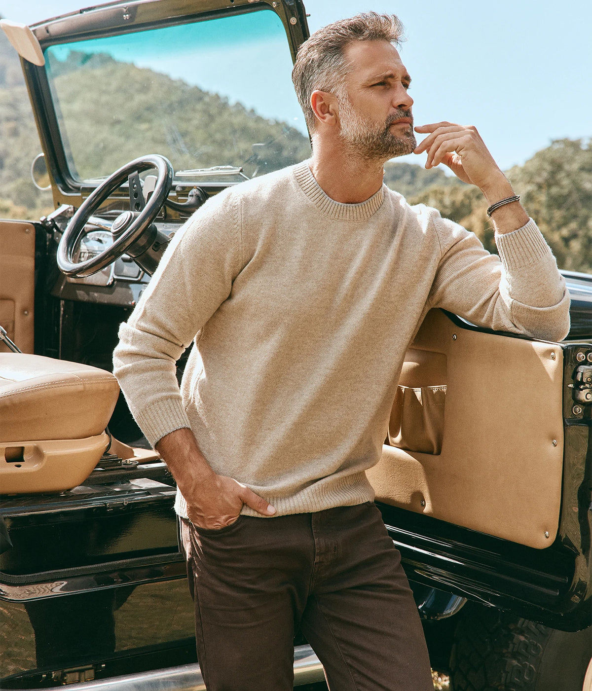 Model wearing the AG Tellis Jean in 7 Yrs Sulfur Oak Barrel with a cream crew-neck knit, leaning back with one hand raised to his chin