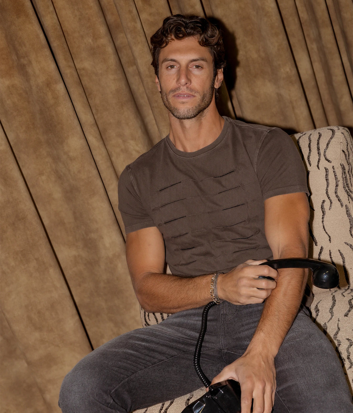 Man wearing John Varvatos Sword Piping Embroidered Tee in Teakwood with denim jeans, seated, front view