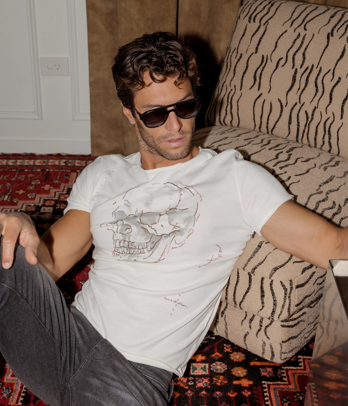 Man wearing John Varvatos Skull Embroidery Raw Edge Tee in Salt with grey jeans and sunglasses, seated on the floor, front view