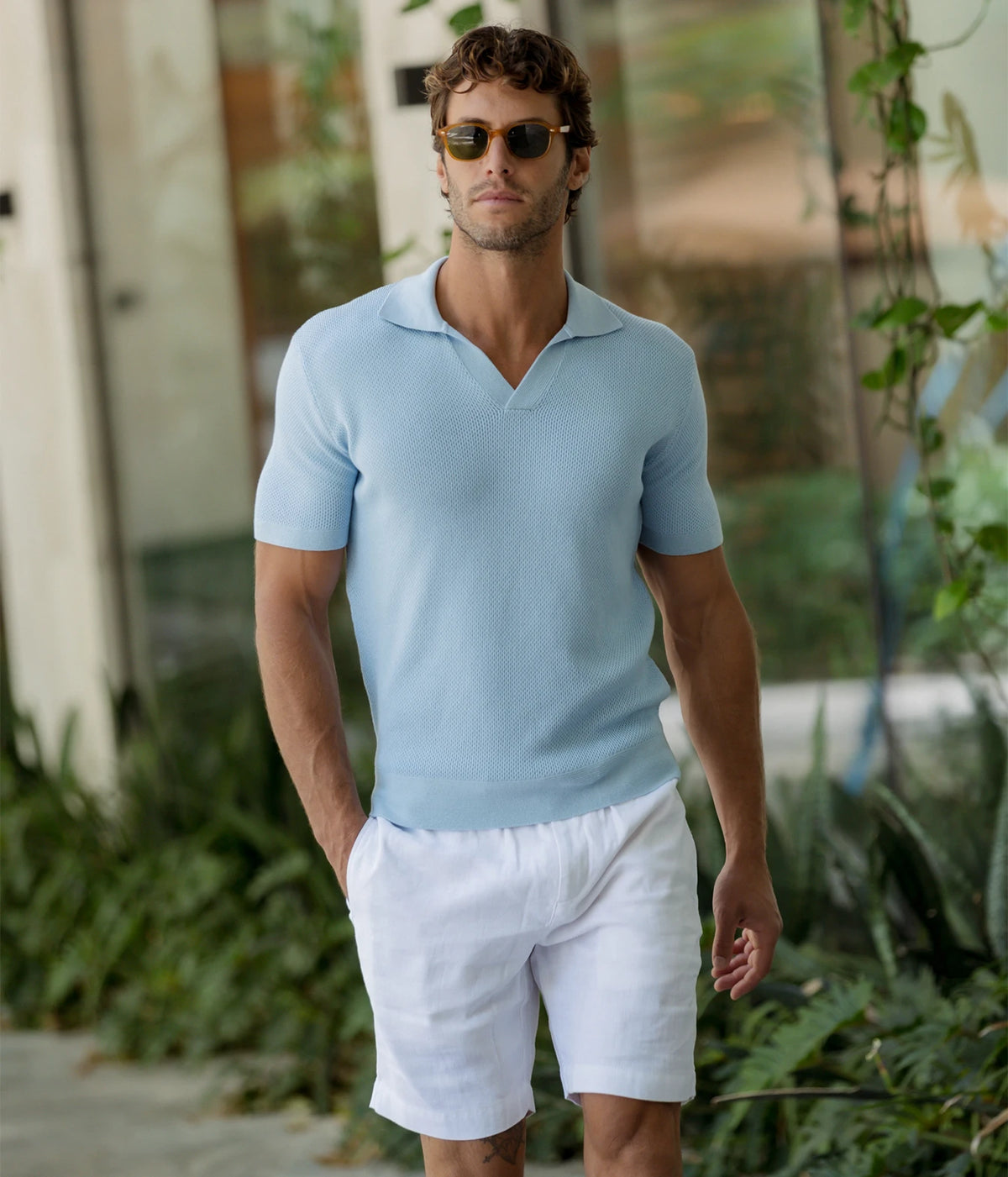 Man wearing Frescobol Carioca Rino Short Sleeve Cotton Polo in Light Denim Blue with white shorts and sunglasses, standing in a garden, front view
