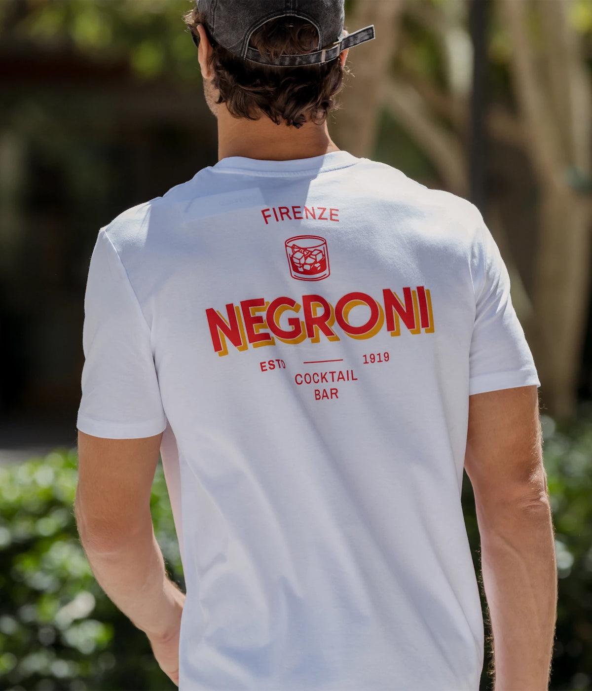 Man wearing The Refined Spirit Negroni Cocktail Bar Tee in White with a cap, outdoors, back view showing the Negroni print