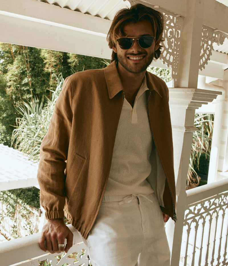 Man wearing a tan Lardini linen jacket with a zip front, elasticated hem and cuffs, and a shirt collar, styled with a white tee and white trousers.