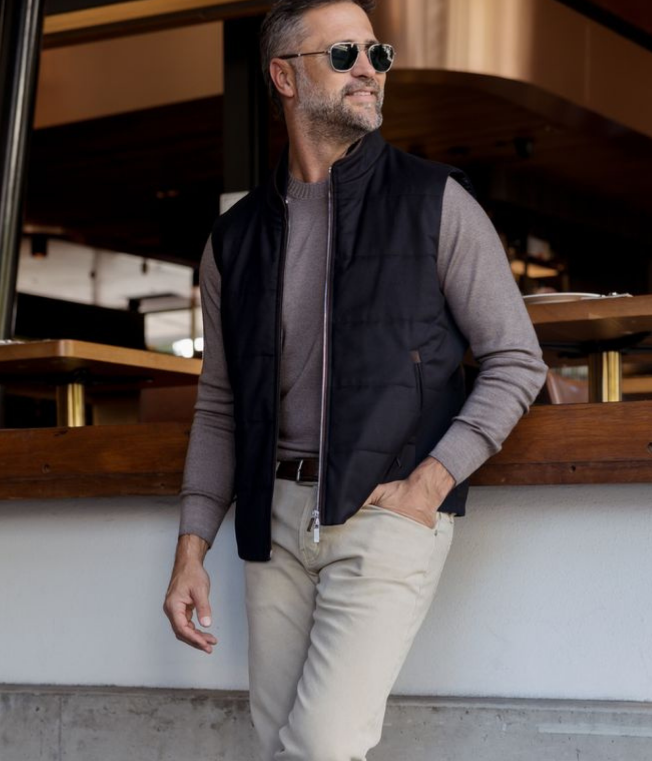 A man wearing a black zip-front puffer vest over a taupe knit sweater, beige trousers, and sunglasses, leaning against a white wall in a modern interior setting.