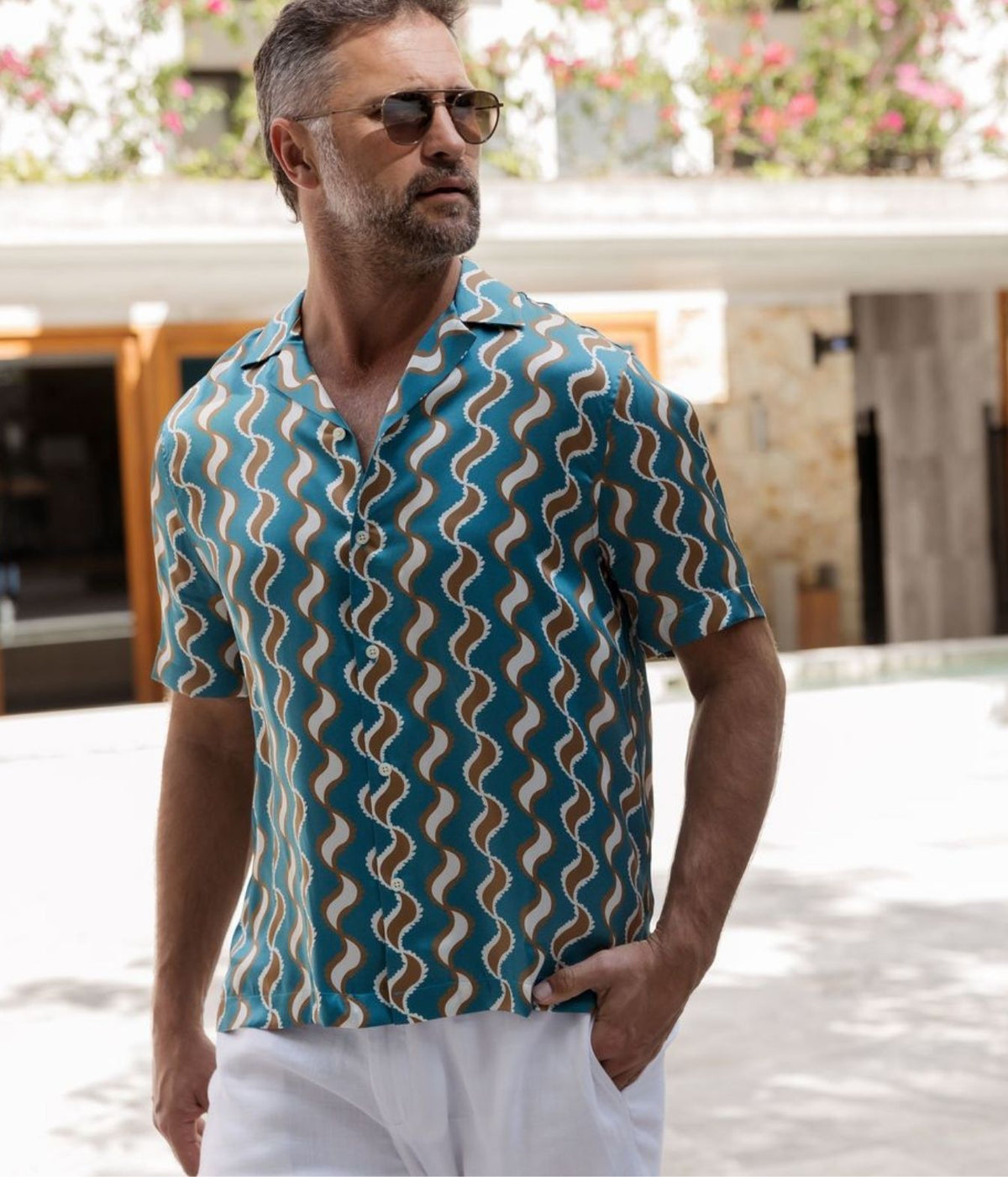 A man wearing a teal short-sleeve button-up shirt with a brown and beige wavy zigzag pattern, paired with white pants and sunglasses, standing outdoors in front of a building.