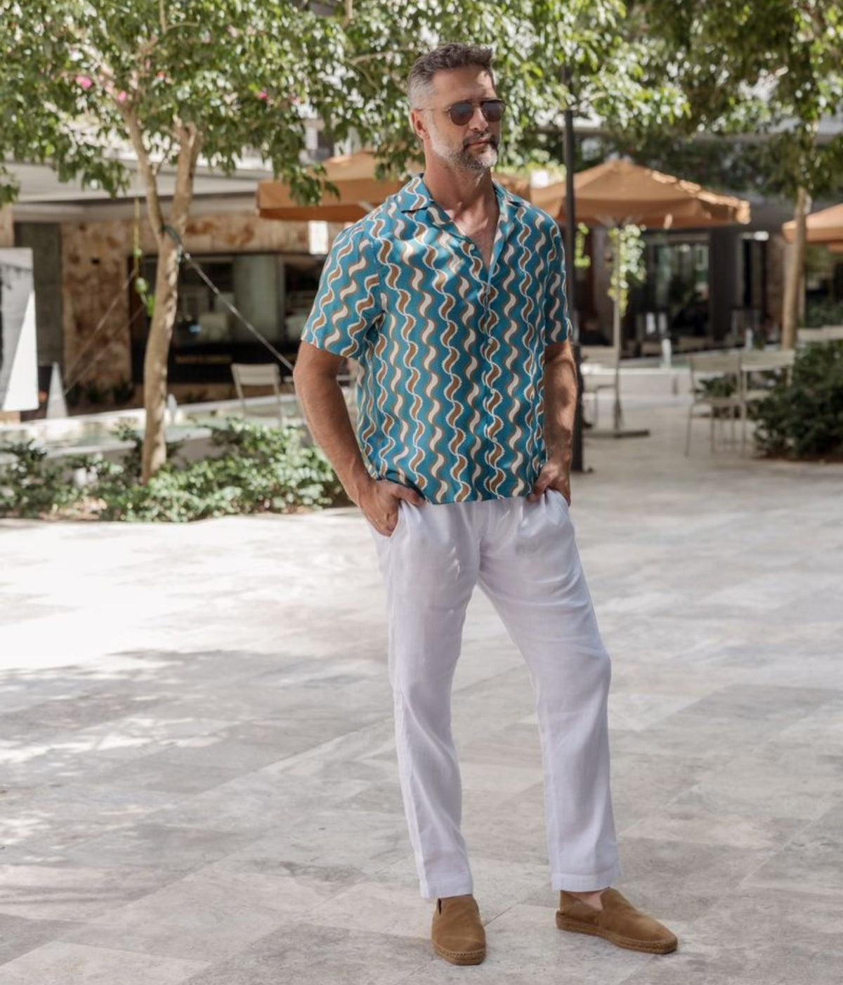 A man wearing a teal and orange zigzag patterned short-sleeve button-down shirt, white trousers, and brown suede loafers, standing in a sunny outdoor patio setting.