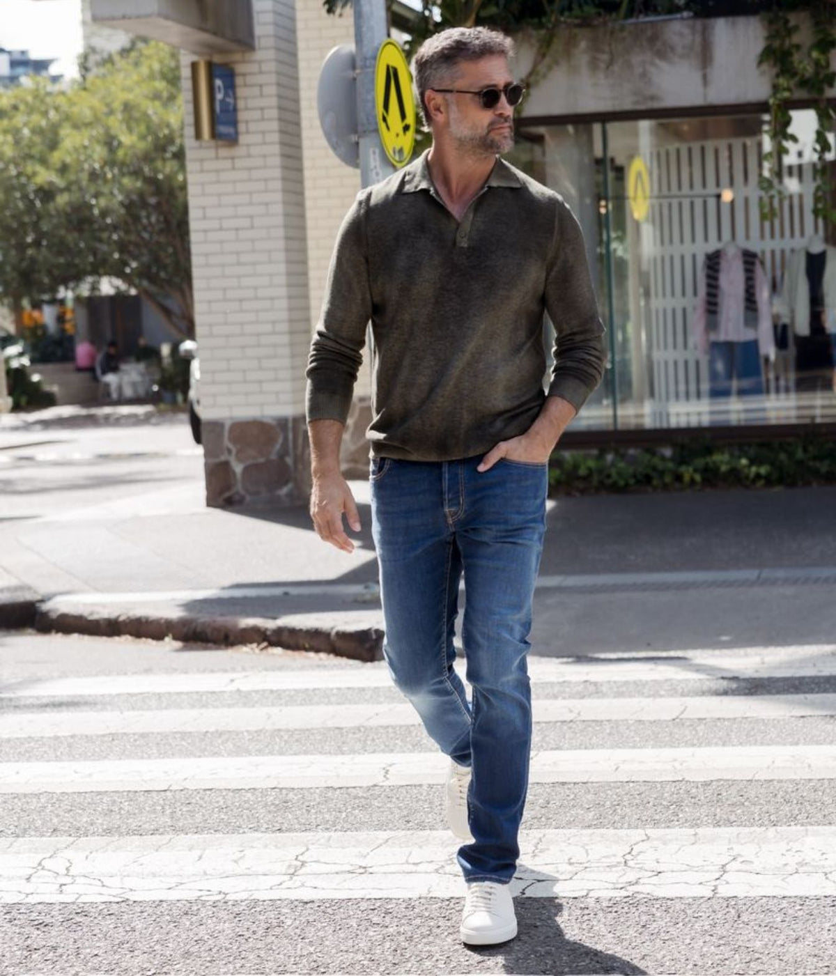 A man wearing a dark olive green long-sleeve polo shirt, blue jeans, and white sneakers walks across a city crosswalk.