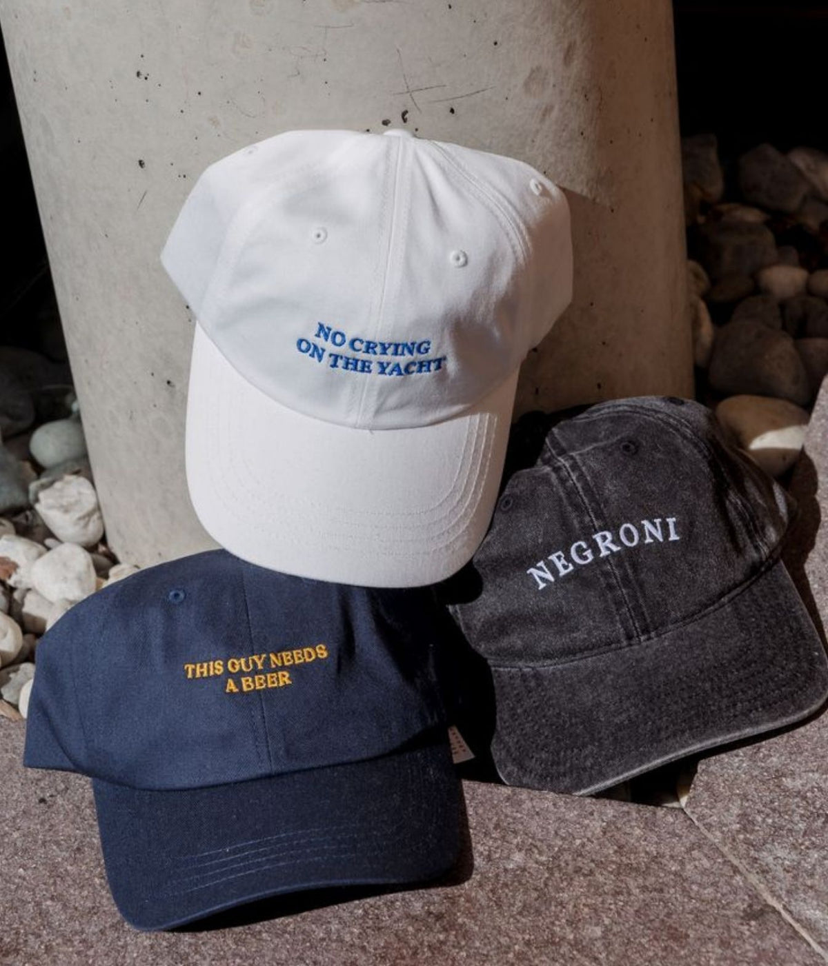 Three embroidered baseball caps arranged on stone pavement in front of a concrete column: a white cap with blue text reading 'NO CRYING ON THE YACHT,' a navy cap with yellow text reading 'THIS OLY NEEDS A BEER,' and a dark charcoal cap with white text reading 'NEGRONI.'.