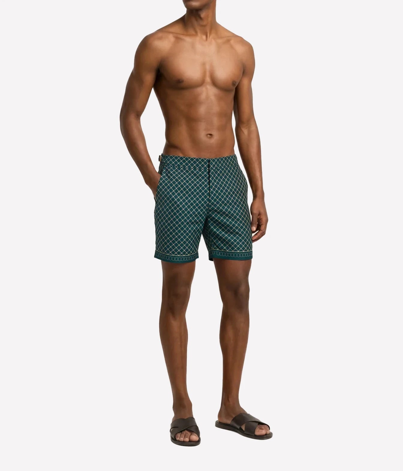 A shirtless male model wearing dark green patterned swim trunks and black slide sandals, standing against a light gray studio background.