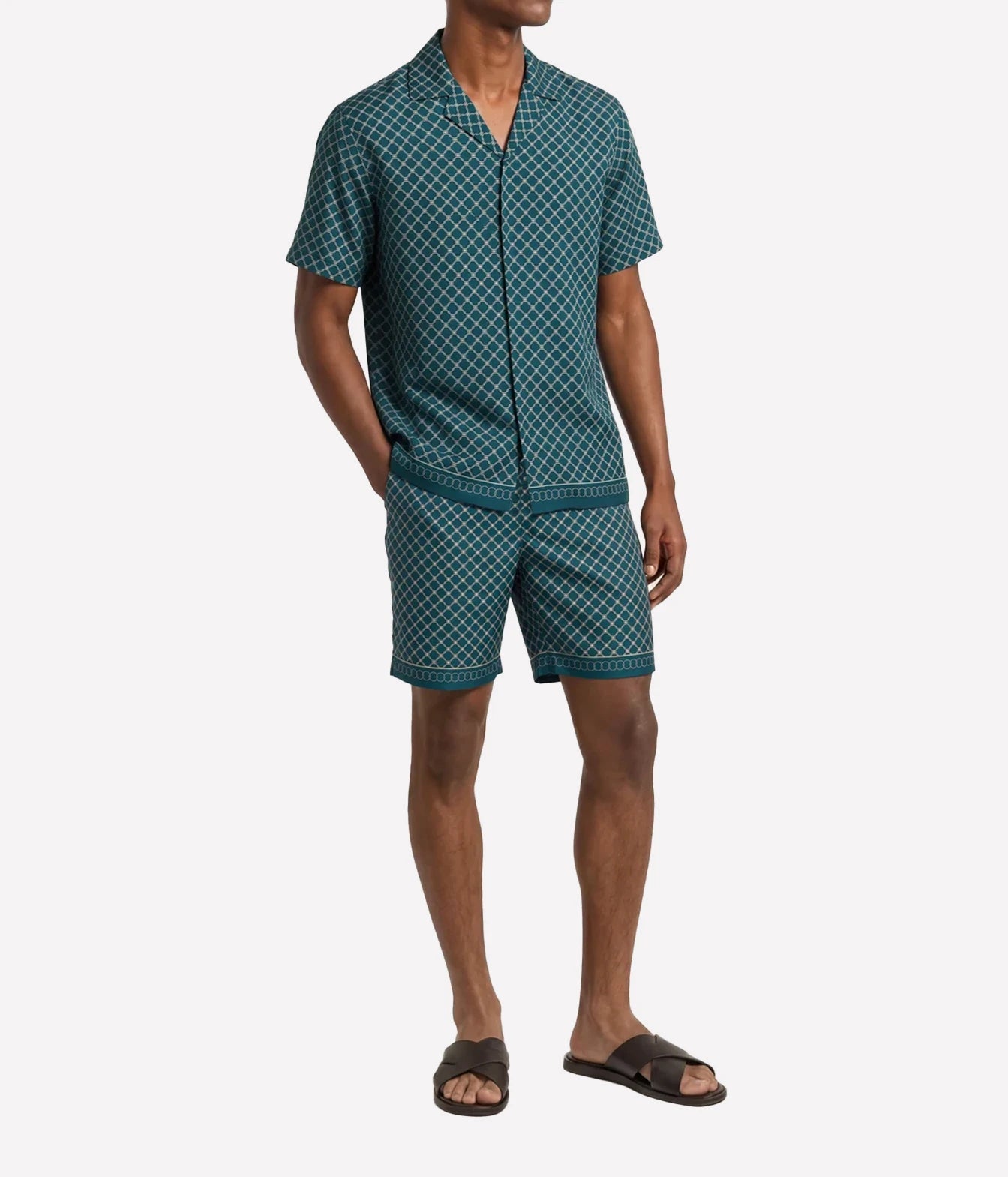 A man modeling a matching short-sleeve button-front pajama shirt and shorts set in a teal and white geometric pattern, paired with dark brown slide sandals, against a light gray studio background.