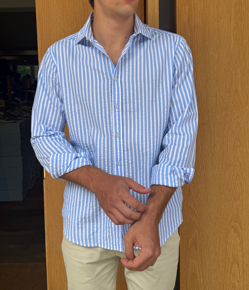 A men's slim-fit striped shirt by Ingram in lightweight blue and white cotton seersucker. Features a button-down front, a crisp collar, and long sleeves with buttoned cuffs. The breathable fabric has a subtle texture, perfect for a polished yet relaxed look.