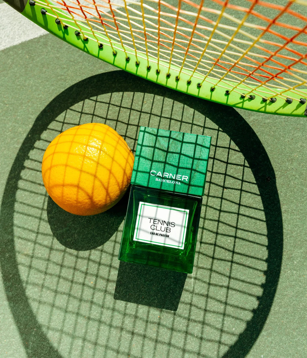 A green tennis-themed perfume bottle labeled 'CARNER TENNIS CLUB' sits on a green court-like surface next to a bright orange, with a tennis racket casting a shadow over the scene.