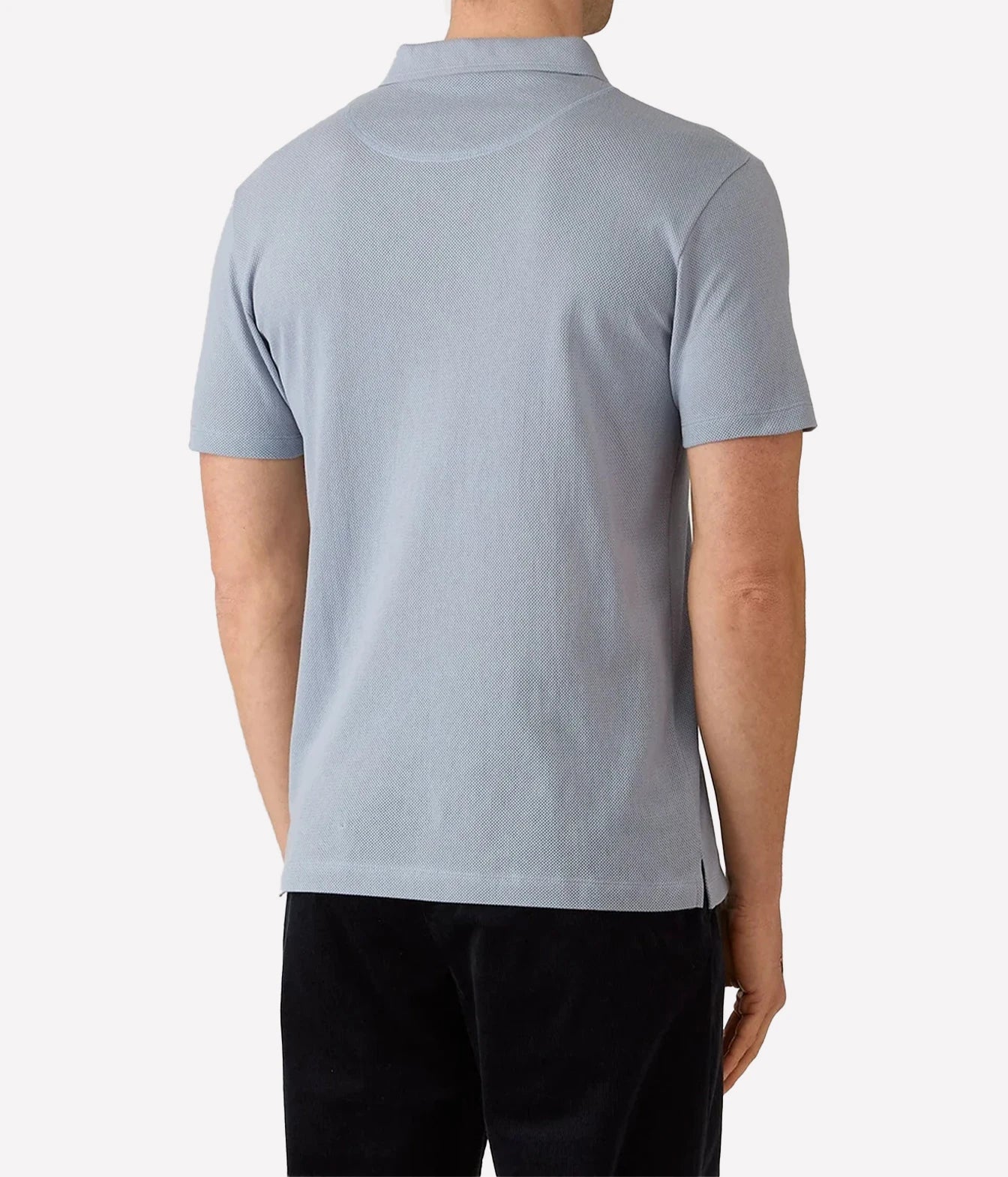 A man viewed from the back wearing a light blue short-sleeve polo shirt and black pants, set against a plain light gray studio background.