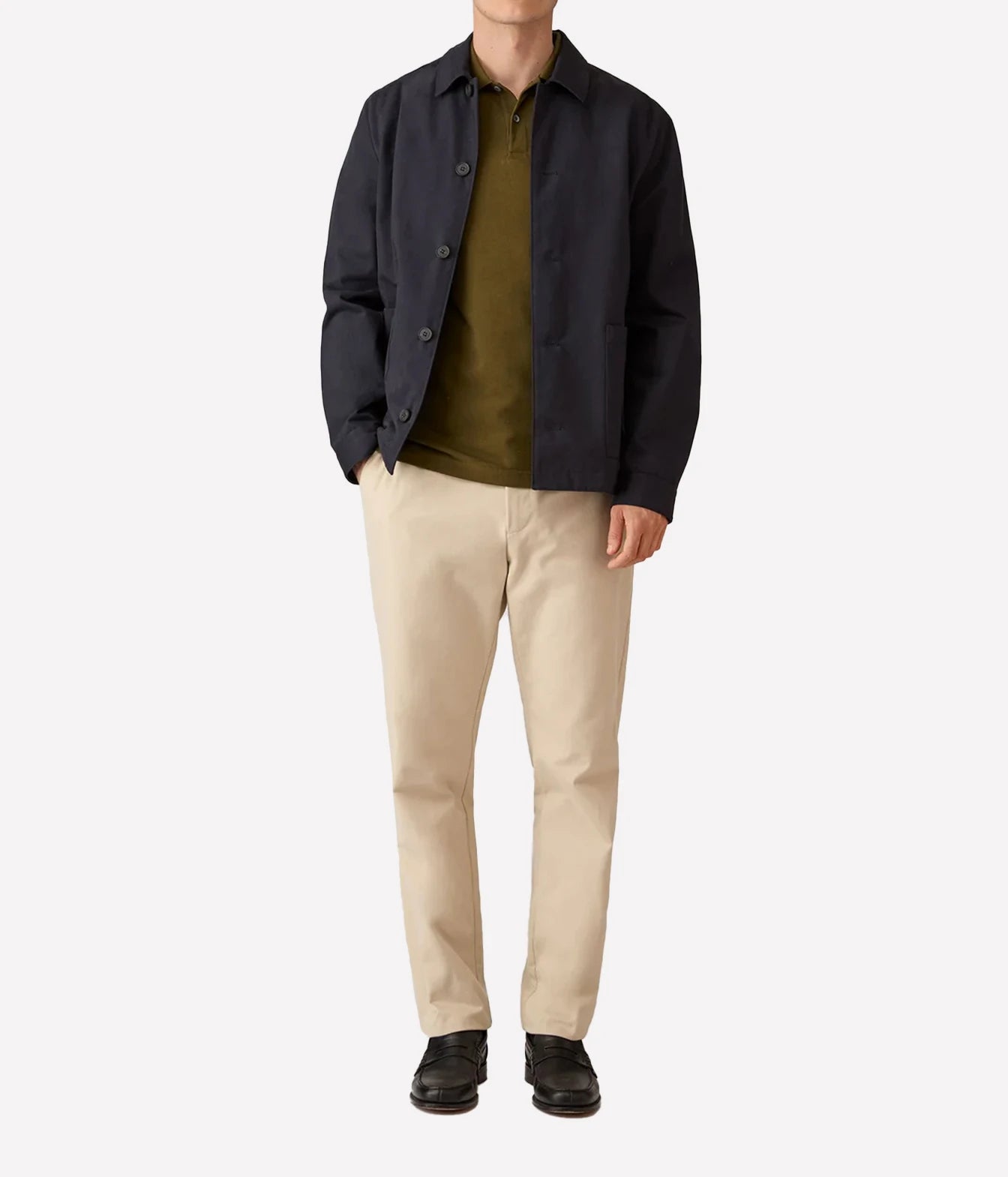 A model wearing a dark navy overshirt, olive green polo shirt, beige trousers, and black shoes, standing against a plain light gray background.