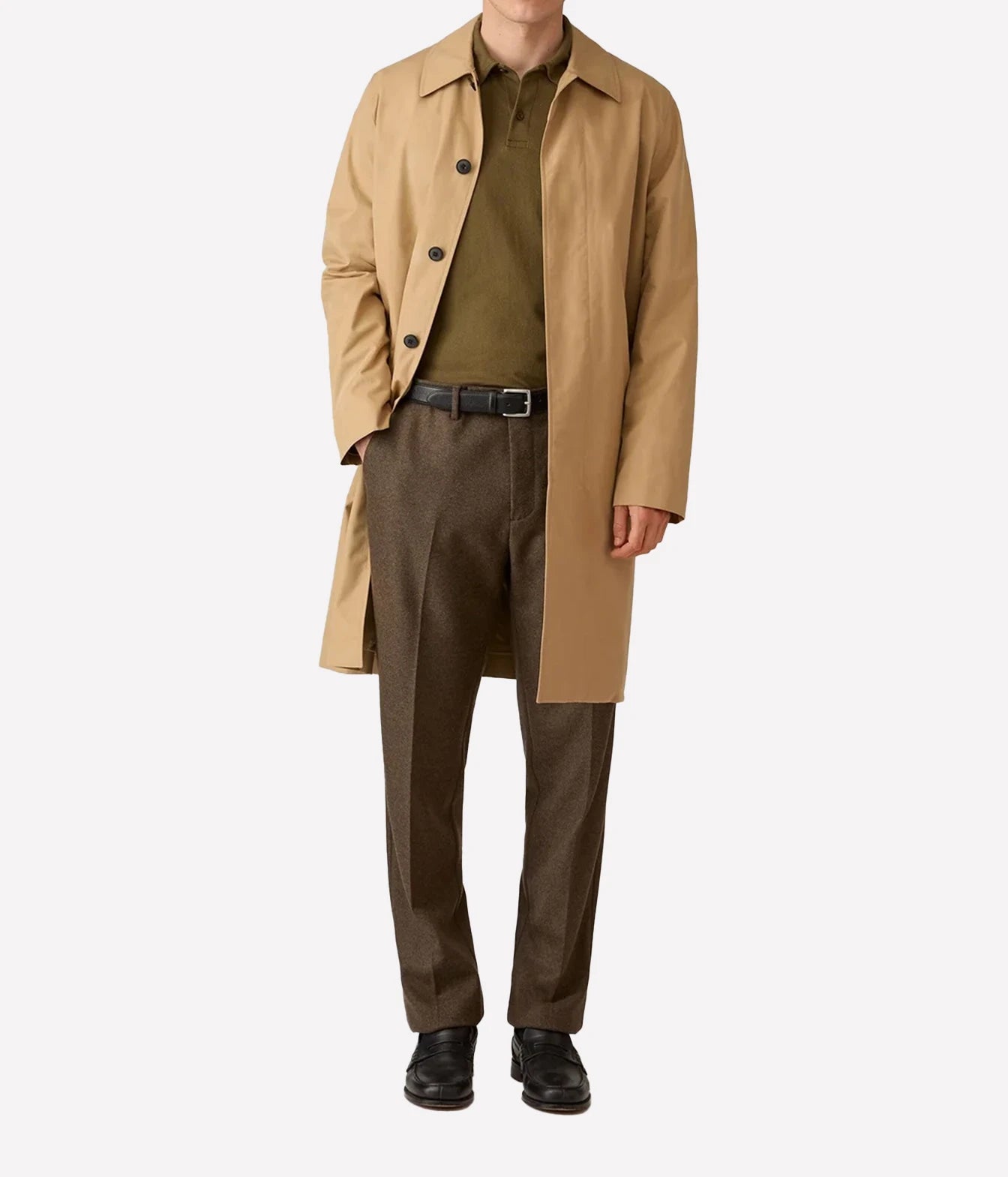 A model wearing a mid-length camel trench coat over an olive green polo shirt, brown textured trousers, a black belt, and black leather shoes, photographed against a solid light gray background.