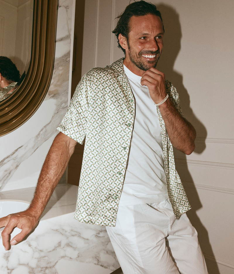 Man wearing a patterned shirt over a white t-shirt and pants in a room with marble flooring.