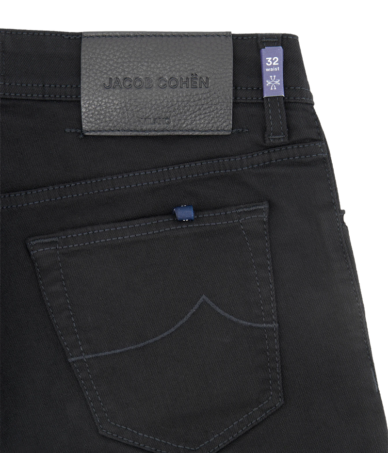 Close-up of the back waistband and pocket of black denim jeans by Jacobs Cohen, showing a dark gray leather brand patch, a blue size tag, and contrast stitching.